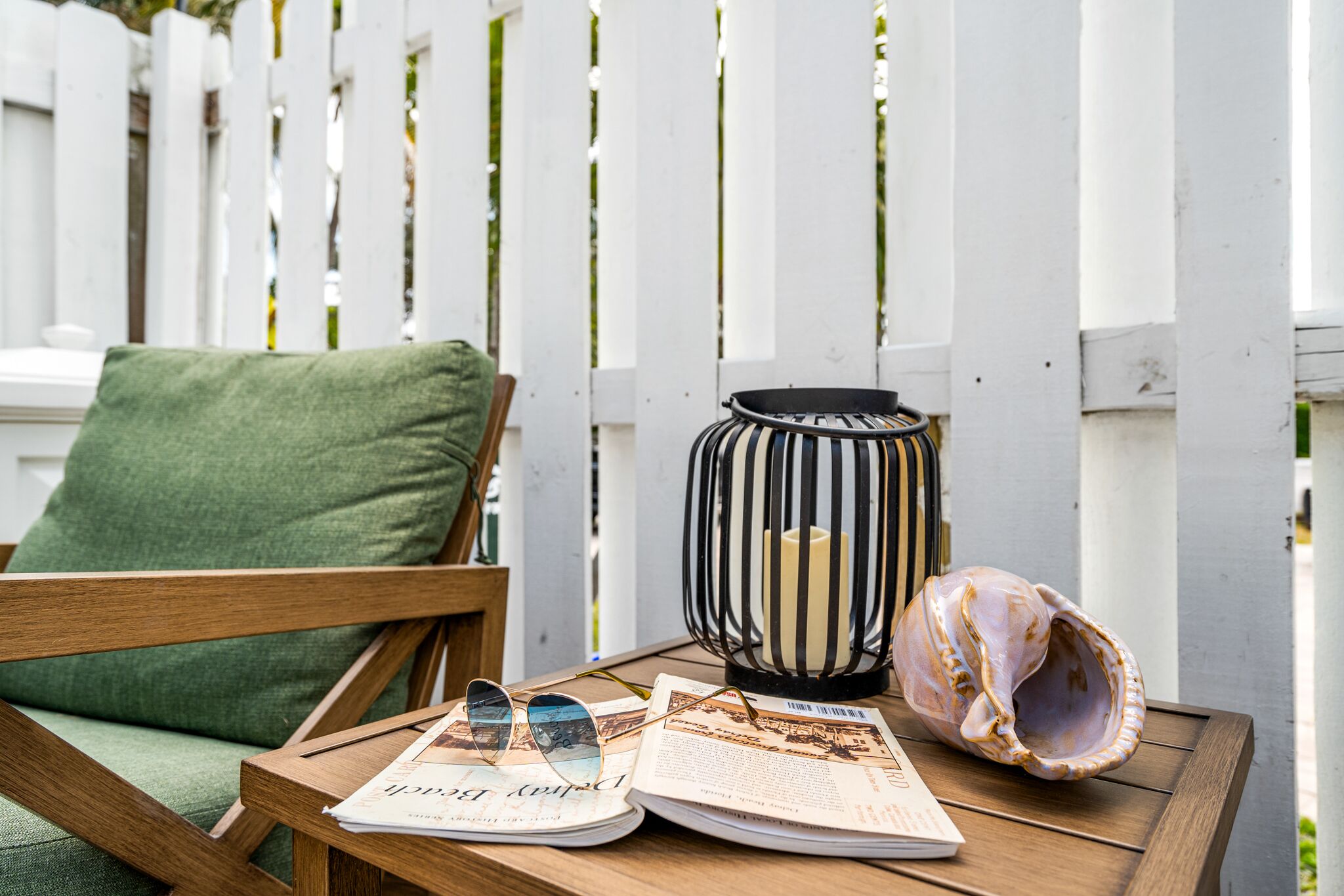 Una mesa de madera con un periódico, un par de gafas de sol, una vela en un farol a rayas y una concha marina.