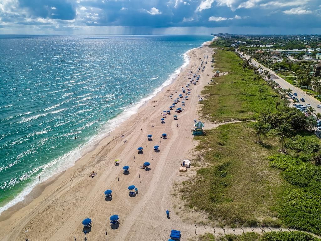 A beach with rows of umbrellas and beach chairs, a road parallel to the beach, and a view of the ocean and distant buildings.
