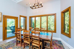 A dining room with a wooden table and chairs, large windows with a view of greenery, and a chandelier.