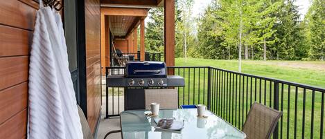 A covered patio with a glass table and chairs, a barbecue grill, and a view of a grassy area with trees.