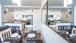 Una cocina moderna con armarios blancos, un microondas y un refrigerador. Hay un comedor con una mesa preparada para dos personas, una botella de vino y un espejo que refleja la cocina.