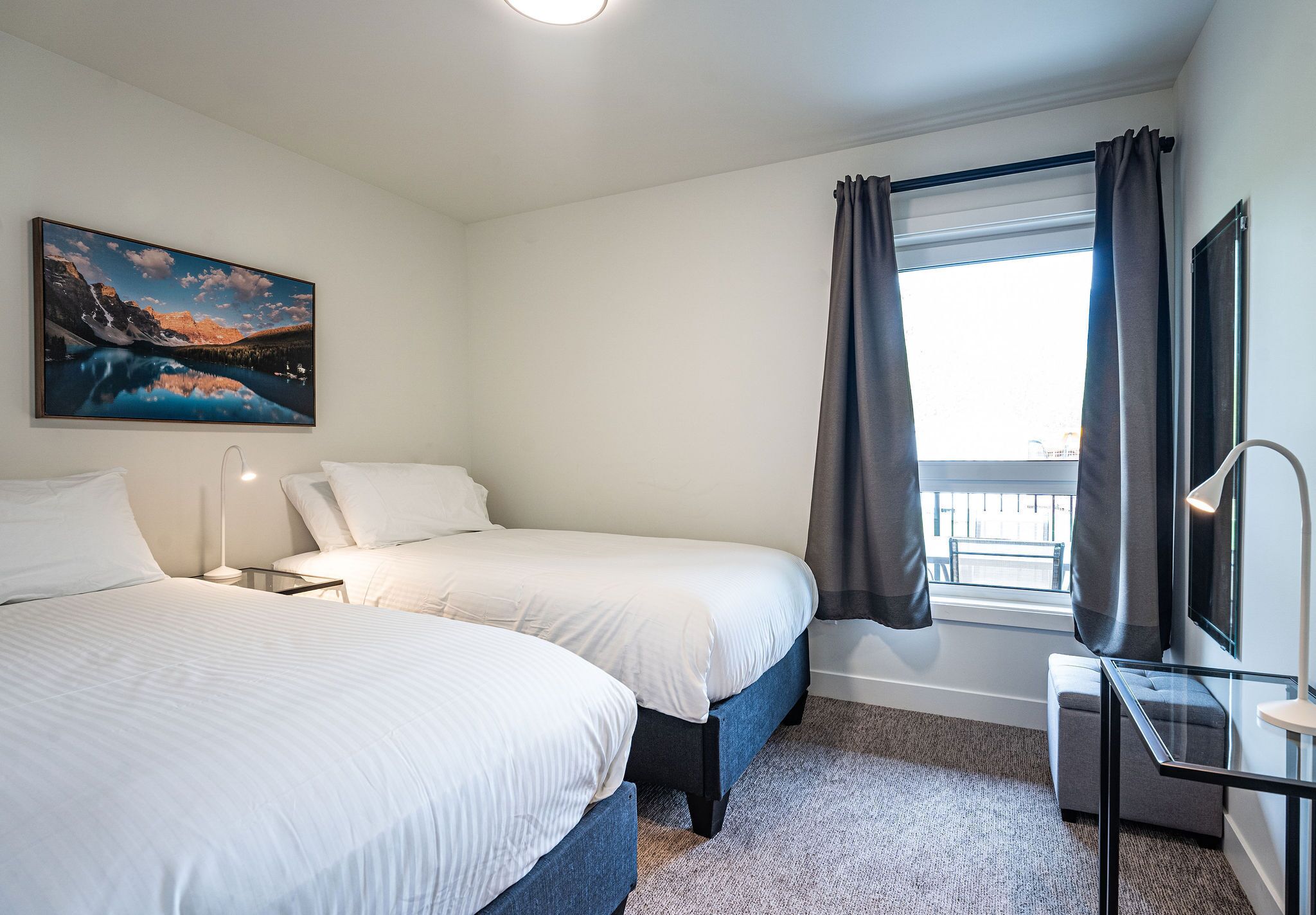 A hotel room with two beds, a desk, a chair, a window with curtains, and a painting on the wall.