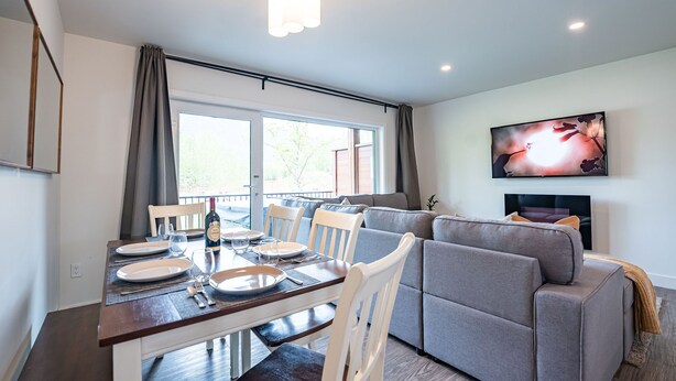 Un comedor con una mesa preparada para cuatro personas, un sofá gris y un televisor montado en la pared.