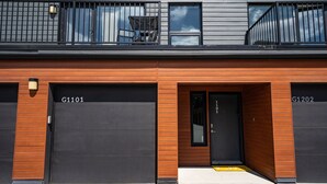 Un edificio moderno con dos puertas de garaje identificadas como G1101 y G1202, una entrada de madera y un balcón con barandilla.
