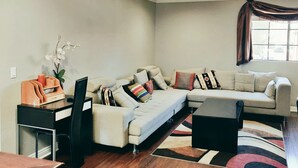 A modern living room with a sectional sofa, a coffee table, and a small side table. There is a window with a curtain and a plant on the side table. - Fully Furnished Style 2 BHK - 1 BATH Apt in San Diego Poway-Miramar with Parking (Poway)