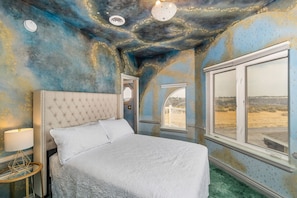 A bedroom with a large bed, a nightstand, a lamp, and a view of the desert through a window.