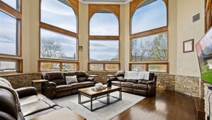 A spacious living room with large arched windows, leather sofas, a coffee table, and a stone accent wall. - Crestview Estate 5 acres-heated pool-5min to strip (Pigeon Forge)
