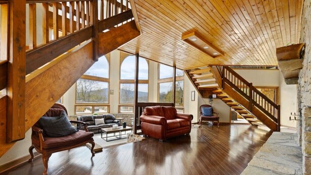 A spacious living area with a wooden staircase, leather sofas, and large windows offering a view of the outdoors. - Crestview Estate 5 acres-heated pool-5min to strip (Pigeon Forge)