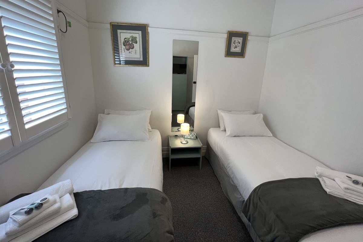 A hotel room with two single beds, a small table with a lamp, a mirror, and two framed pictures on the wall.