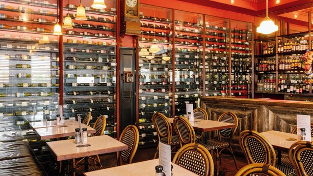 Un restaurant avec plusieurs tables et chaises, un présentoir à bouteilles de vin en arrière-plan et une horloge au mur.