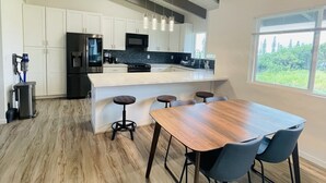 A modern kitchen with white cabinets, a stainless steel refrigerator, a microwave, and a large island with bar stools. - Big Island Green Sands House (Naalehu)