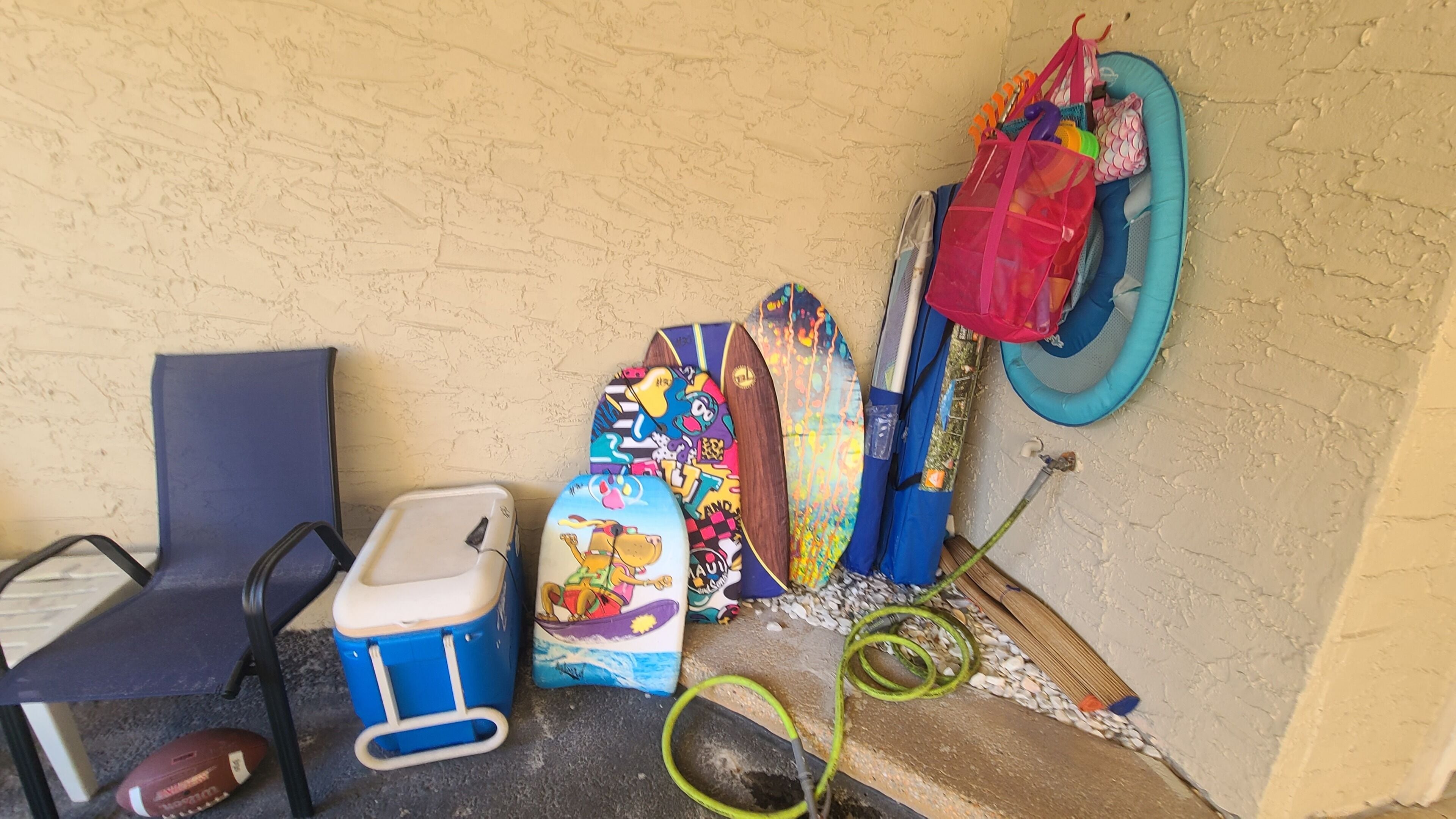 A corner with a blue chair, a blue cooler, colorful bags, a blue inflatable ring, and a green hose.