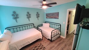 Une chambre avec deux lits simples, un ventilateur de plafond, un tableau accroché au mur et un téléviseur fixé au mur.