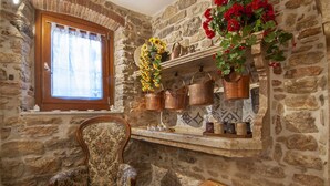 Interior rústico de pared de piedra con una silla de madera, una ventana con cortina y una repisa con ollas de cobre y flores rojas.