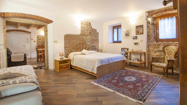 Un dormitorio con pared de piedra, cama con cabecera de madera, mesita de noche, una silla, una pequeña mesa con lámpara y alfombra.
