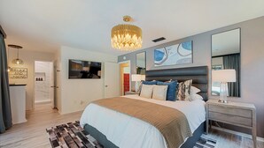 A modern bedroom with a large bed, bedside lamps, a chandelier, a mirror, and a wall art piece.