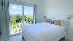 A bedroom with a bed, a ceiling fan, a window with curtains, and a view of greenery outside. - Ocean View 1 BR w/Office, Rooftop and Gardens (Playas del Coco)