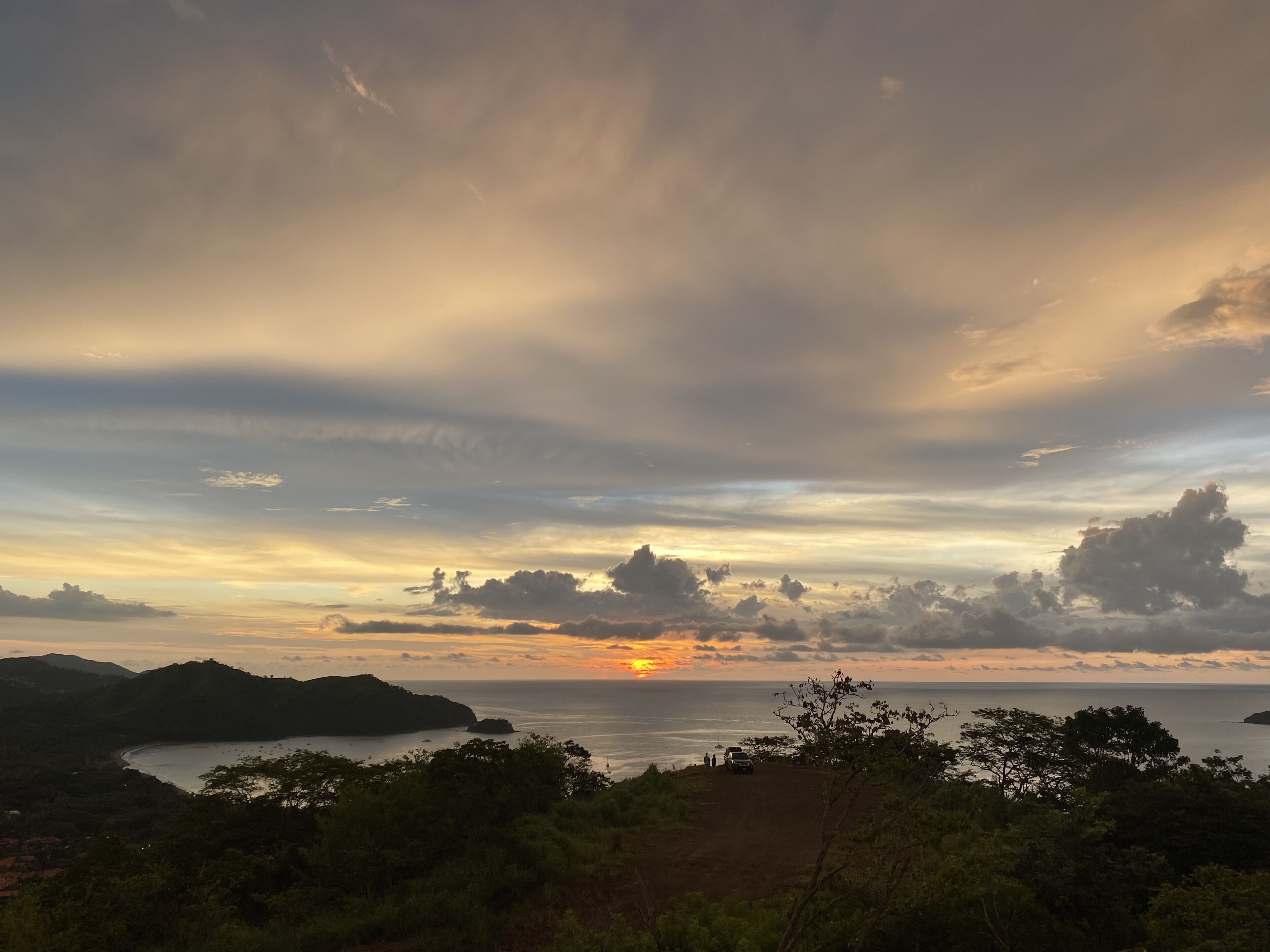 A coastal landscape with a sunset, hills, and a road with a vehicle.