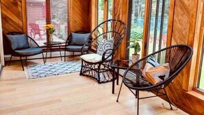 A cozy room with wooden walls and large windows, furnished with black wire chairs and a small table with a vase of flowers. - Spacious Riverfront Getaway in the Heart of the White Mountains (Carroll)