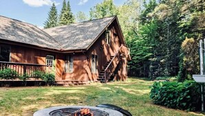 A rustic cabin with a stone fire pit in the foreground, surrounded by a well-maintained garden and tall trees. - Spacious Riverfront Getaway in the Heart of the White Mountains (Carroll)