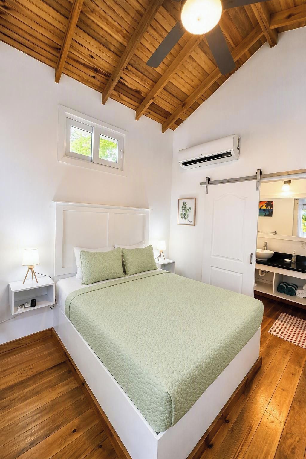A bedroom with a wooden ceiling, a bed with a green bedspread, a bedside table with a lamp, and an air conditioning unit.