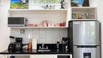 Uma cozinha moderna com geladeira de aço inoxidável, micro-ondas e cafeteira. Há armários, pia e uma janela com vista para o verde.