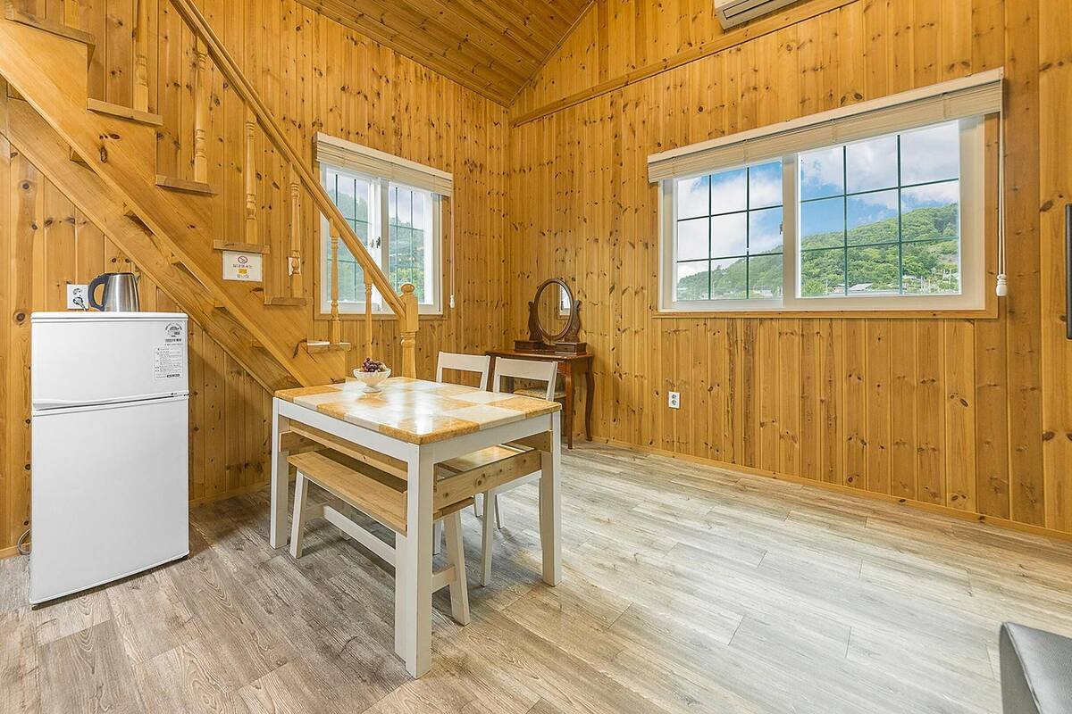 A wooden-paneled room with a kitchen area, including a refrigerator, a table with chairs, and a window with a view of greenery.