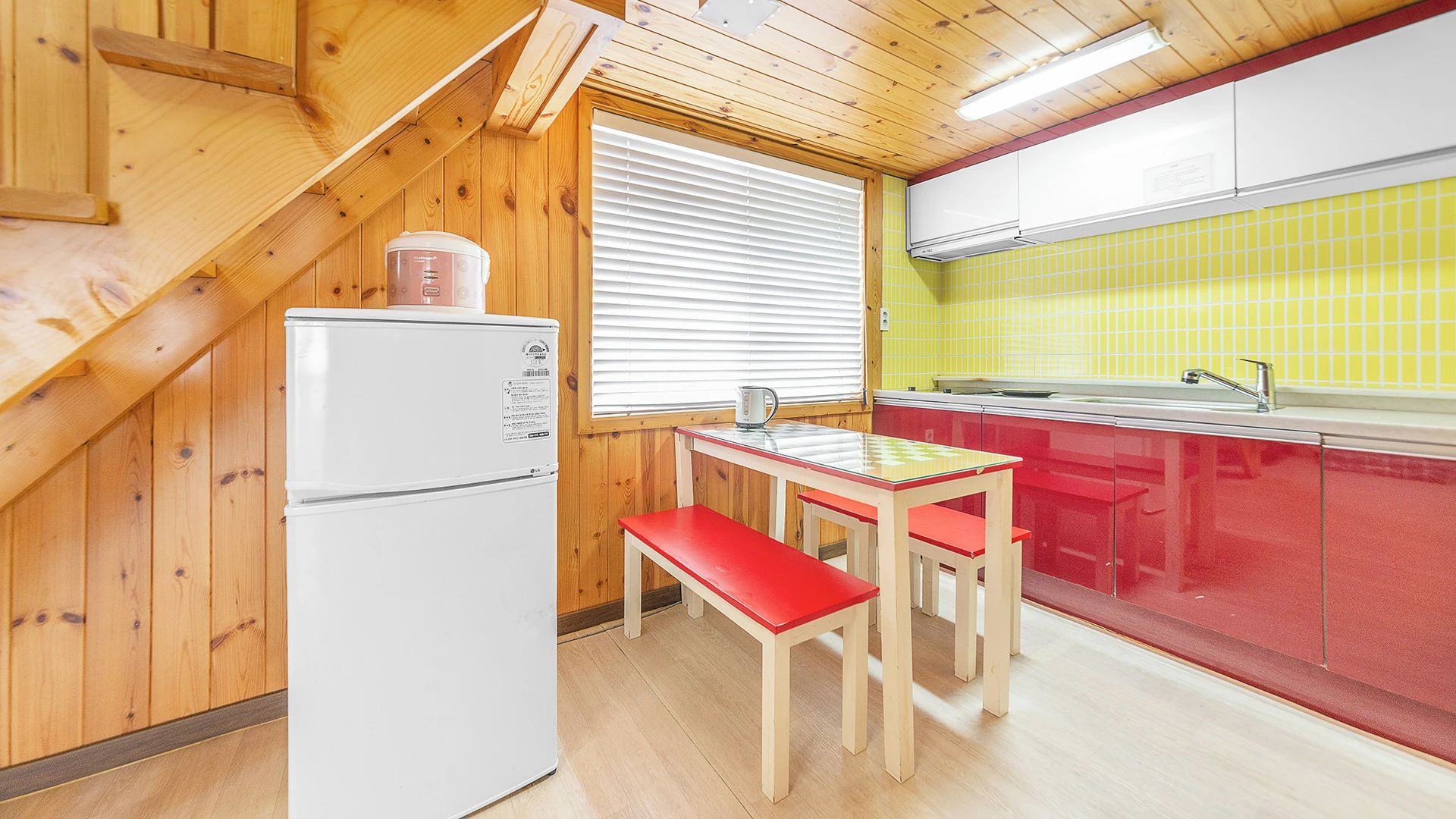 Une cuisine de taille réduite, équipée d’un réfrigérateur, d’une table avec deux chaises et d’une fenêtre avec des stores.