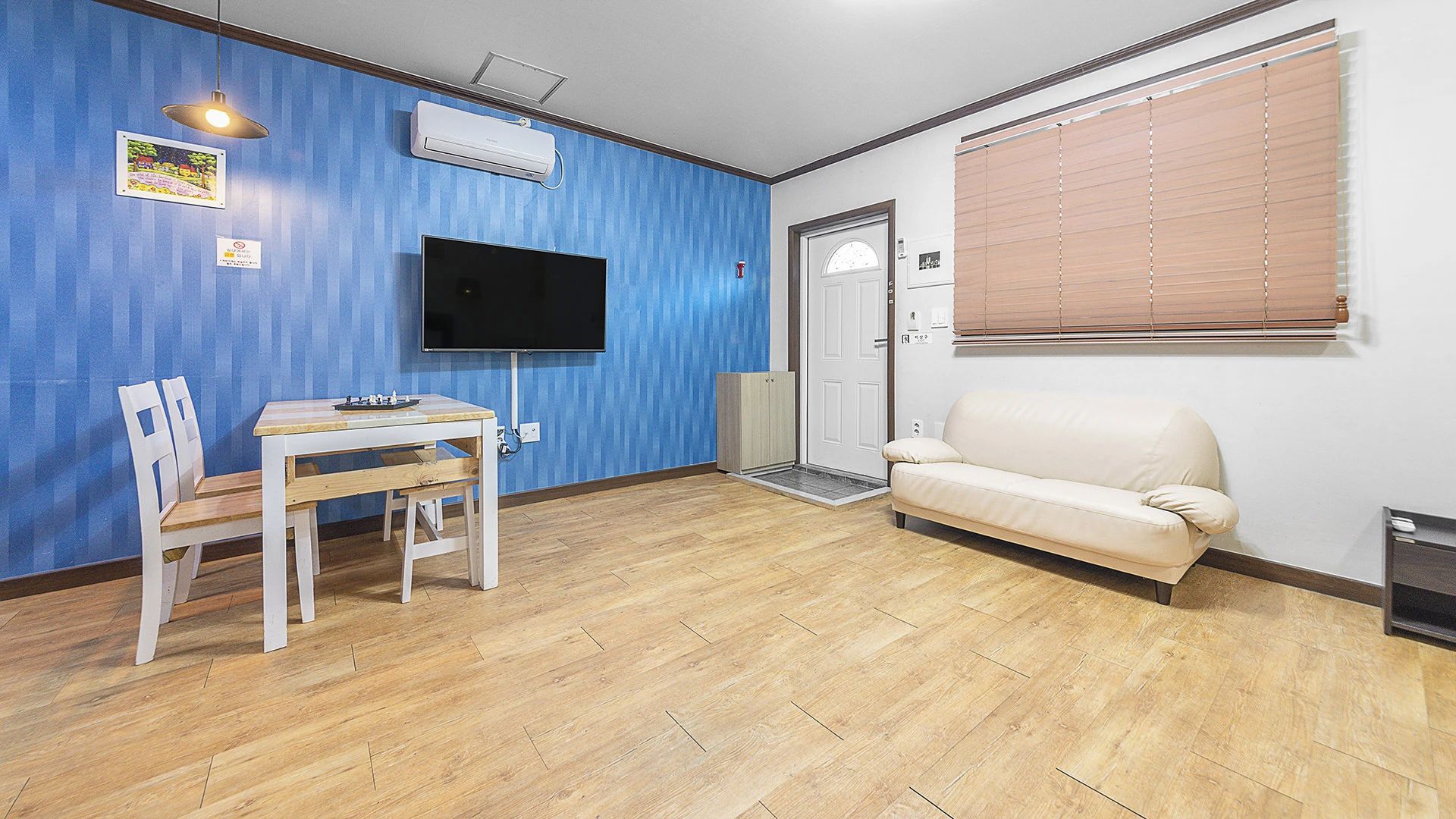 A room with a wooden floor, a white sofa, a TV mounted on the wall, and a door with a window.