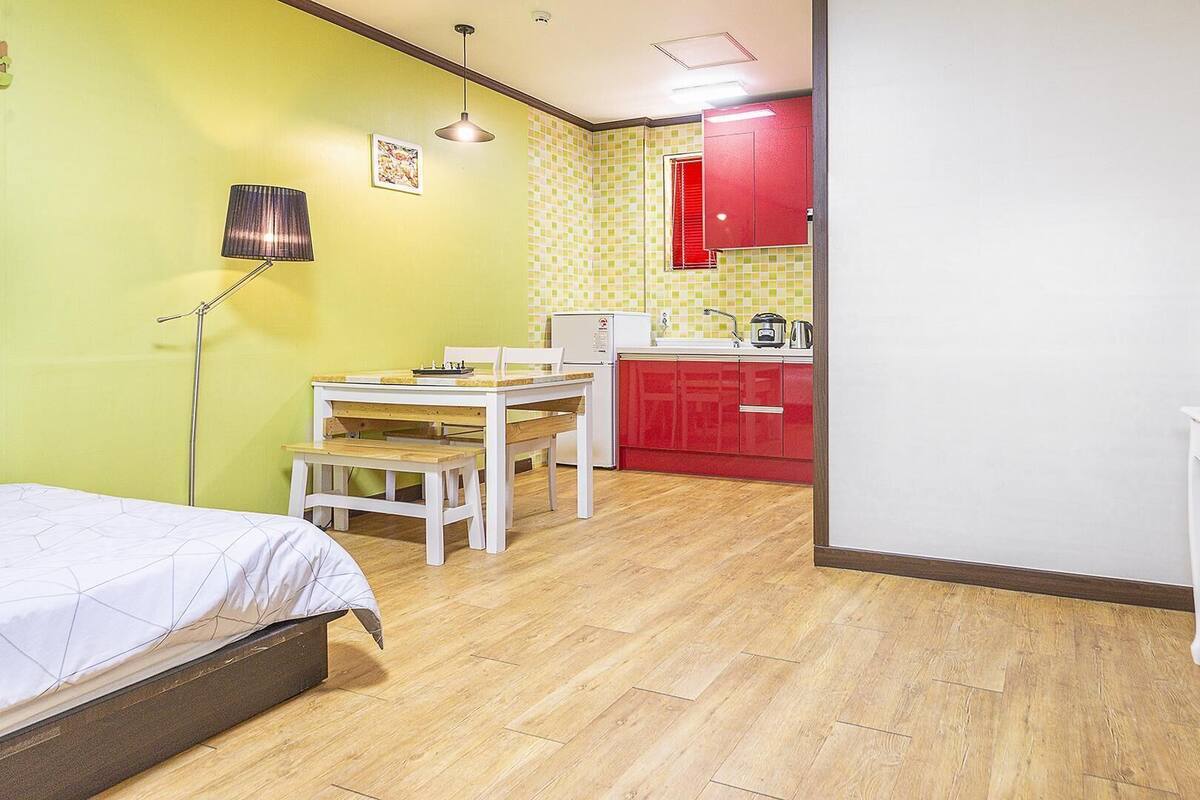 A compact room with a bed, a dining table, a small kitchen area, and a mirror.