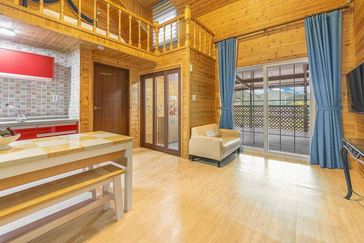 A wooden cabin interior with a kitchen area, a dining table, a sofa, and a large window with curtains.