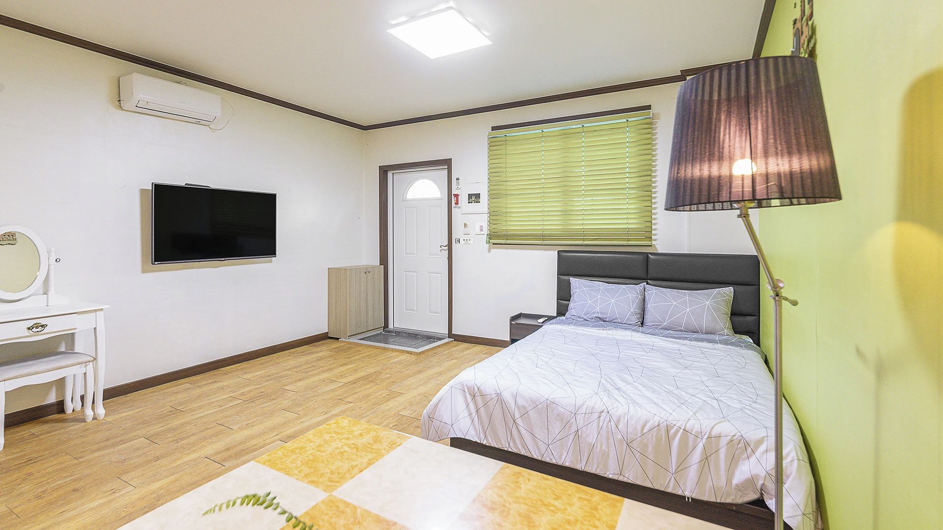 A bedroom with a bed, a desk with a mirror, a television, and a window with blinds.