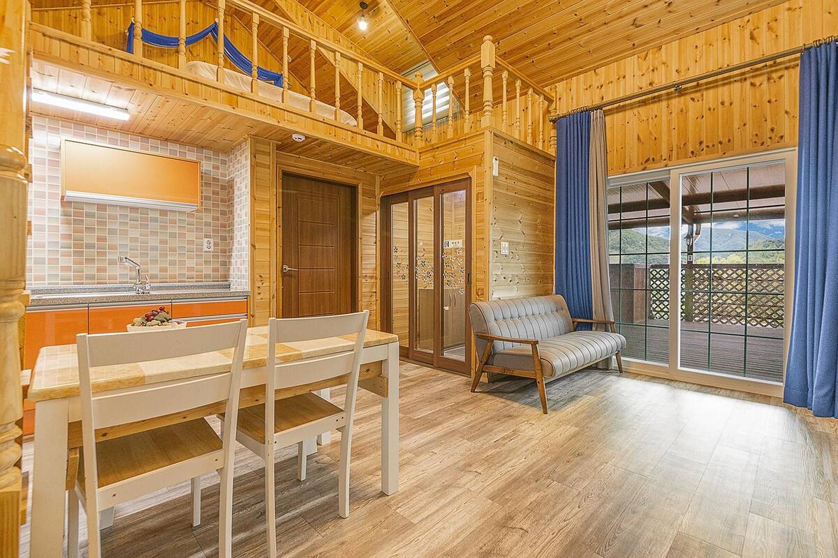 A wooden cabin interior with a dining area, a sofa, and a large window with curtains.