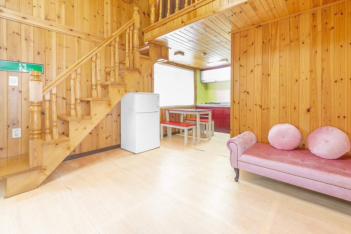 A wooden interior with a staircase, a white refrigerator, a pink sofa with round cushions, and a dining area with red chairs.