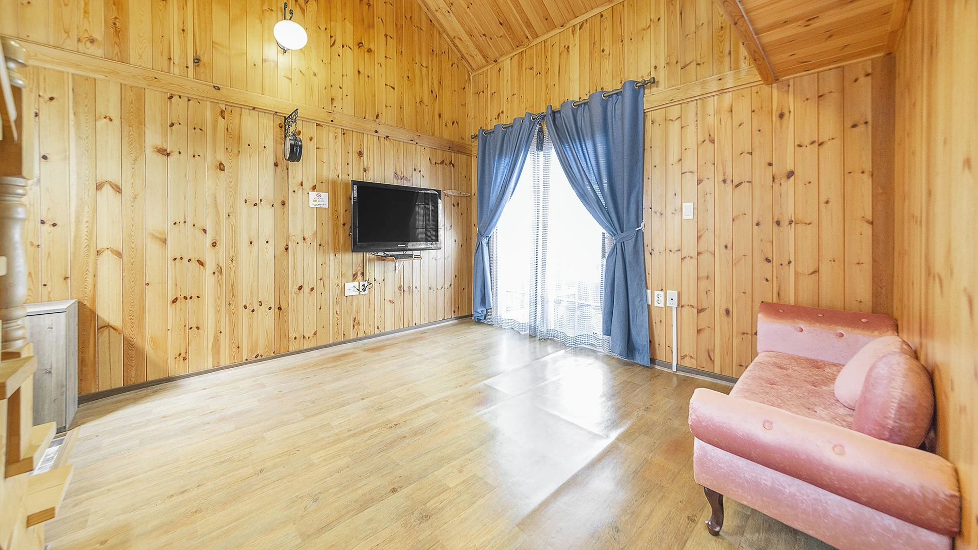 A room with wooden walls, a flat-screen TV mounted on the wall, a pink sofa, and a window with blue curtains.