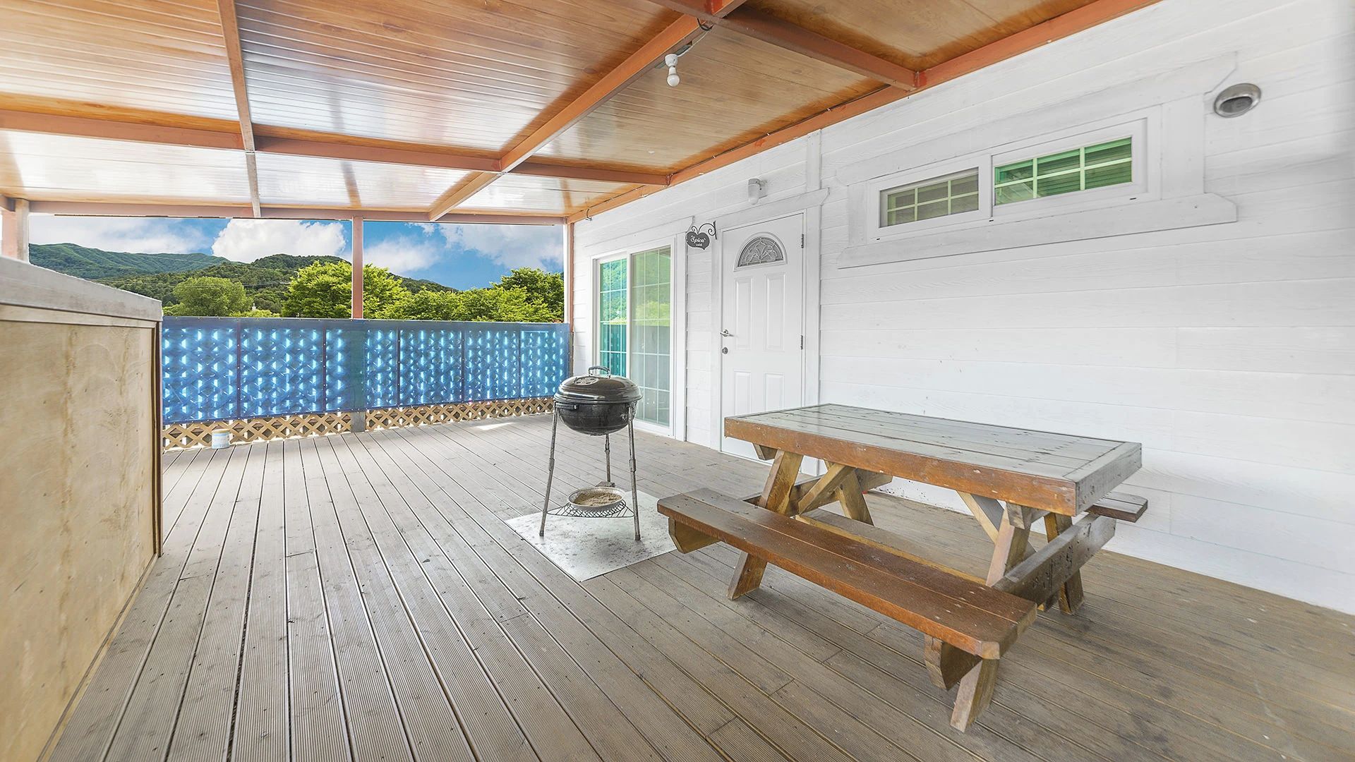 Een terras buiten met een barbecue en houten tafel, omgeven door een witte muur en uitzicht op groen.