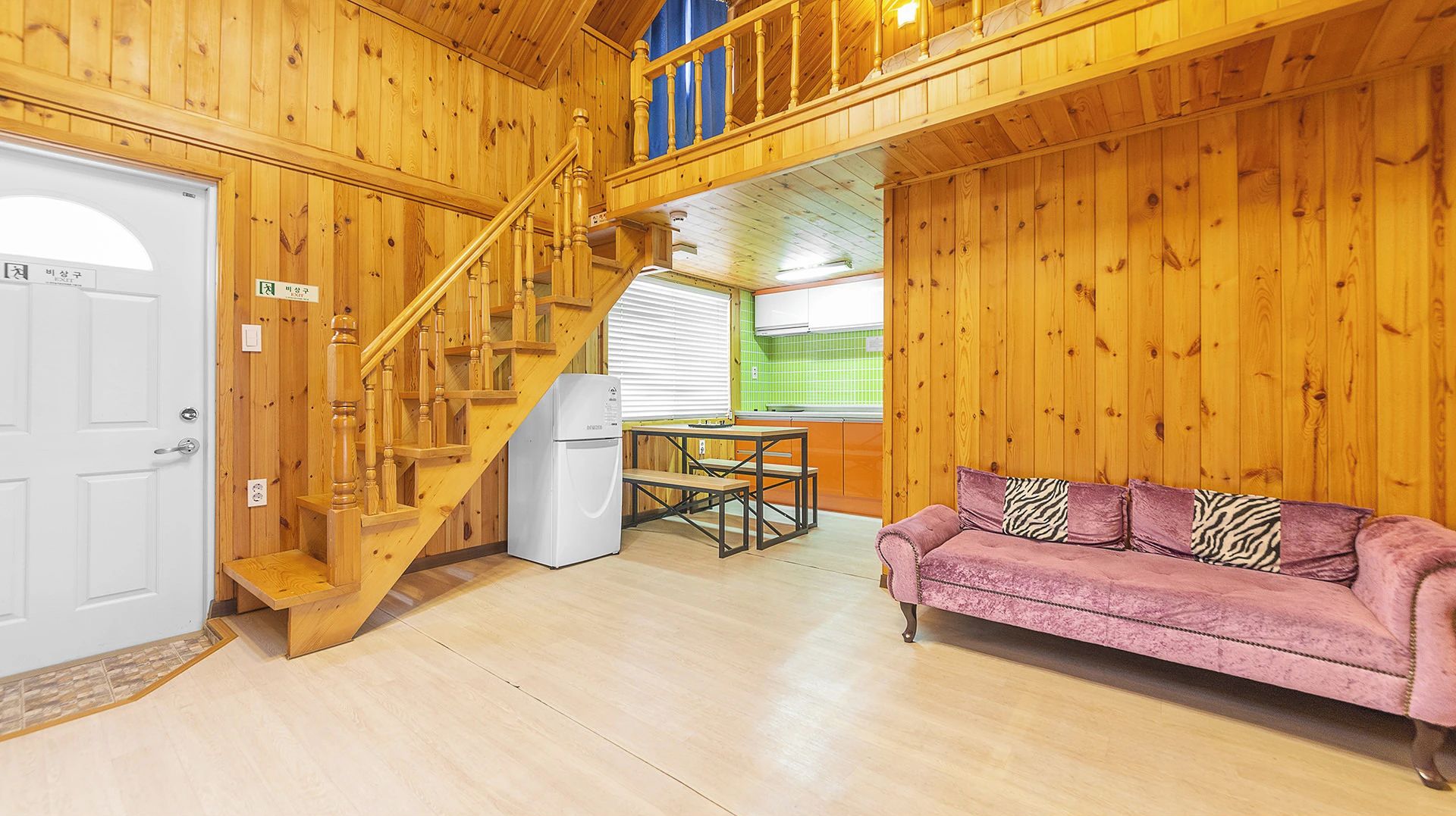 Un intérieur en bois avec un escalier, un réfrigérateur blanc, un canapé rose et une table à manger.