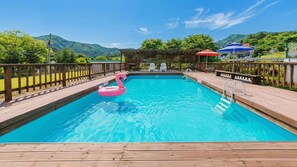 Una piscina rectangular amb un trampolí de fusta, un flamac amb color rosa i una pèrgola de fusta amb cadires.