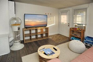 Ein Wohnzimmer mit Fernseher auf einem Holzständer, einer weißen Spielzeug-Gumballmaschine, einem Korbwäge und einem runden Holztisch mit Zeitschrift und Fernbedienung.