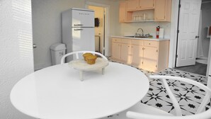 Une cuisine compacte avec un réfrigérateur blanc, un évier et un plan de travail. On y trouve une table ronde blanche, avec une chaise placée devant.