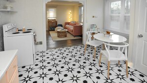 Une cuisine équipée d’une cuisinière, de meubles blancs et d’un sol orné d’un motif en étoile. On y trouve également un coin repas avec une table et des chaises blanches, ainsi qu’un salon avec un canapé et une lampe.