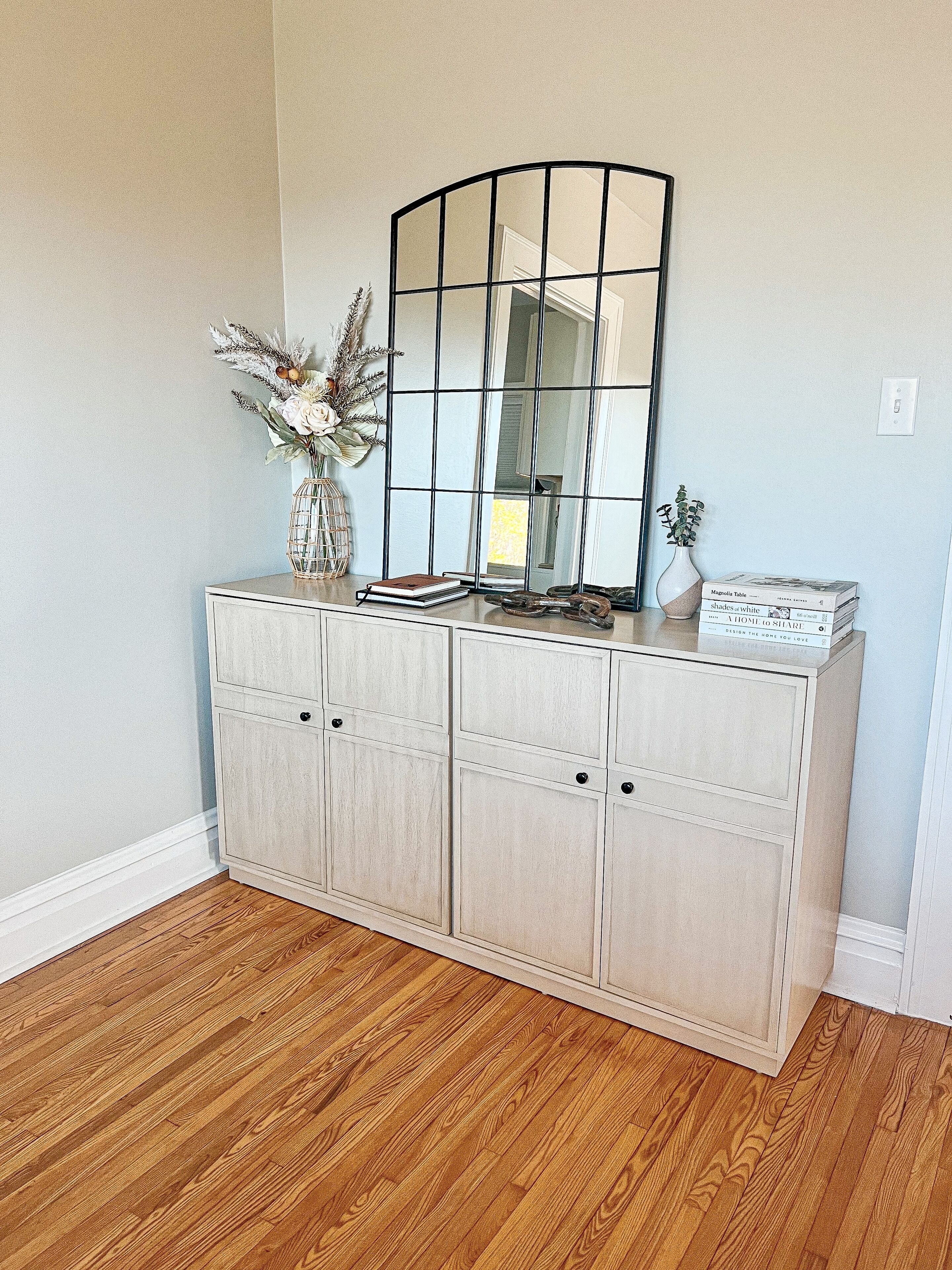A white wooden cabinet with a mirror above it, a vase with dried flowers, and a stack of books.
