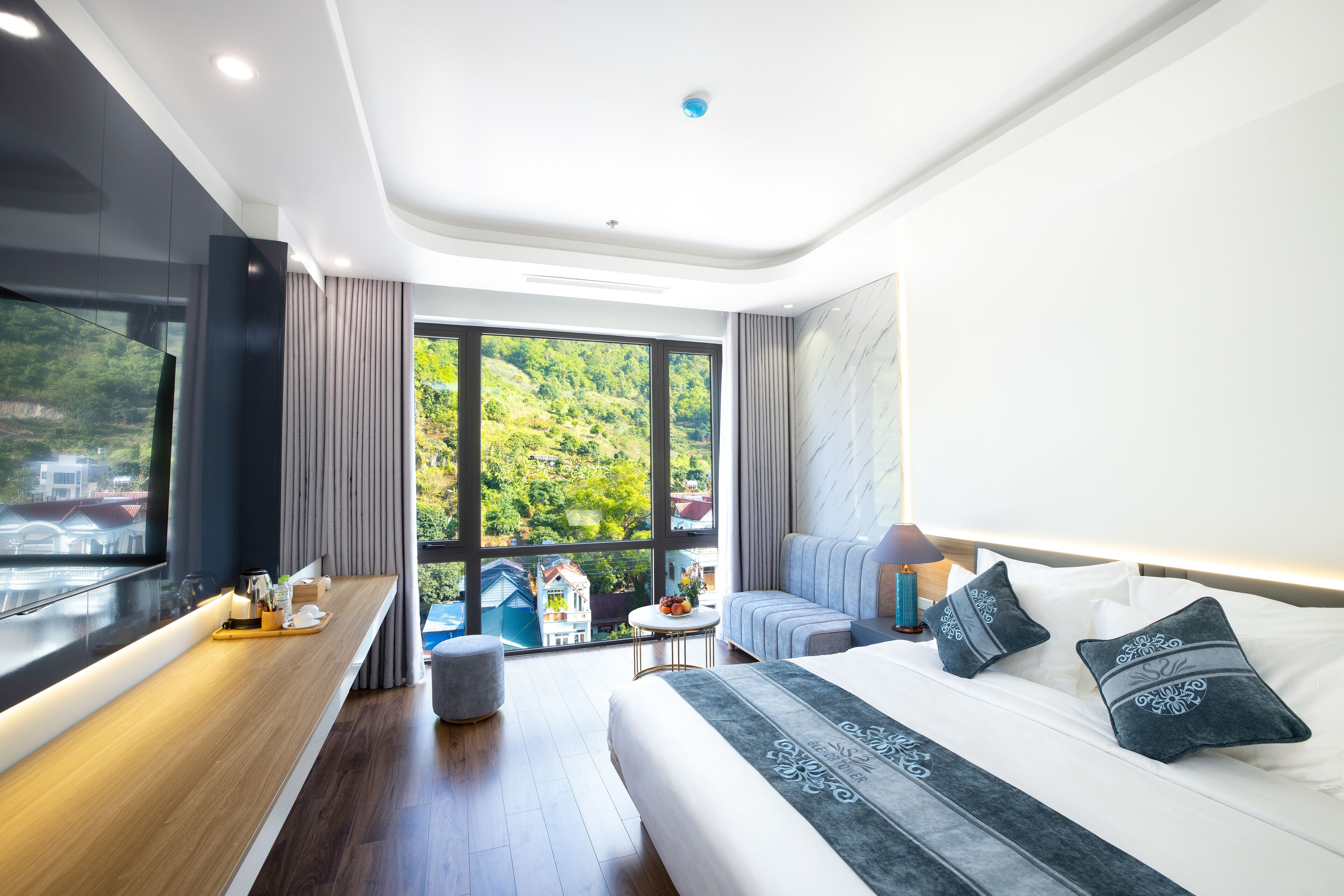 Een moderne hotelkamer met een groot raam, een bed met decoratieve kussens, een nachtkastje met een lamp en een klein rond tafeltje met fruit en een vaas.