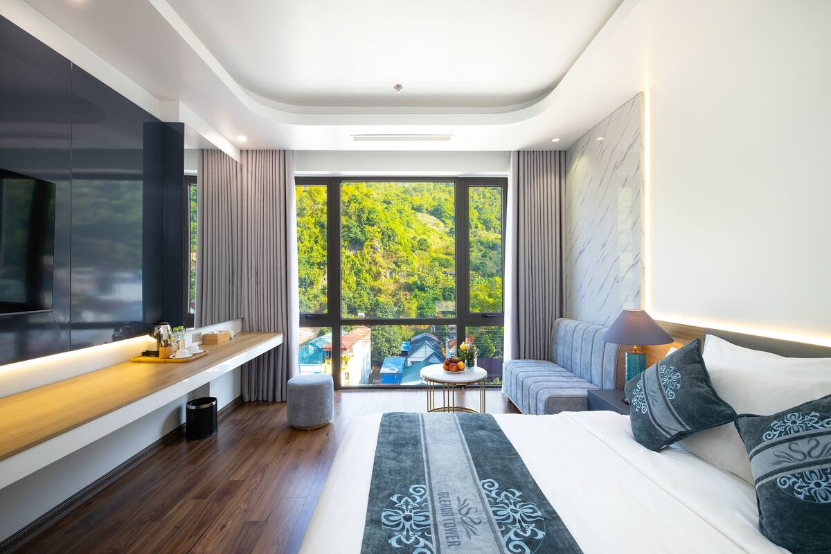 A modern hotel room with a large bed, a TV, a wooden bench, and a view of greenery outside.