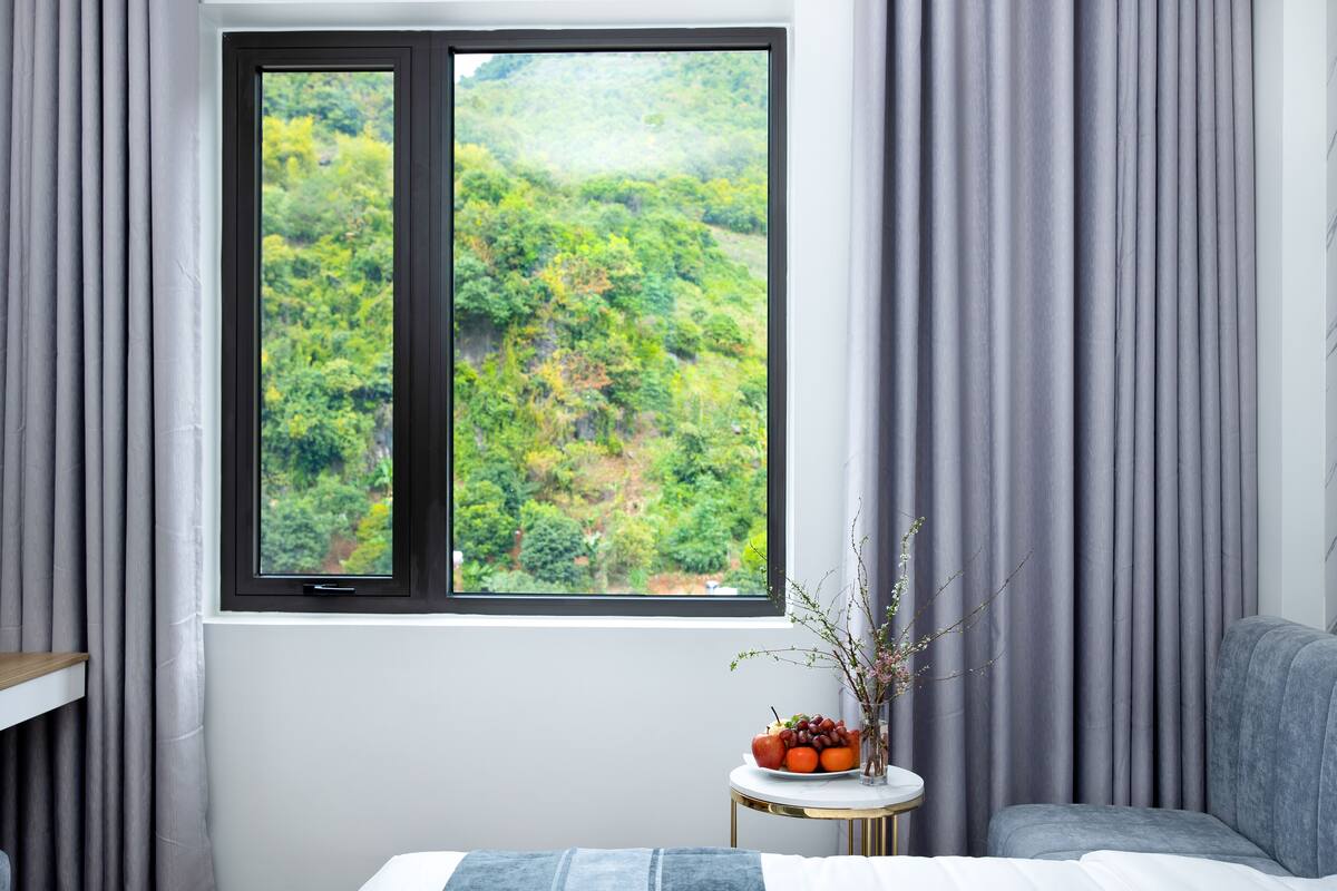 A room with a large window, a bed, a bedside table with a vase of flowers, and a grey sofa.