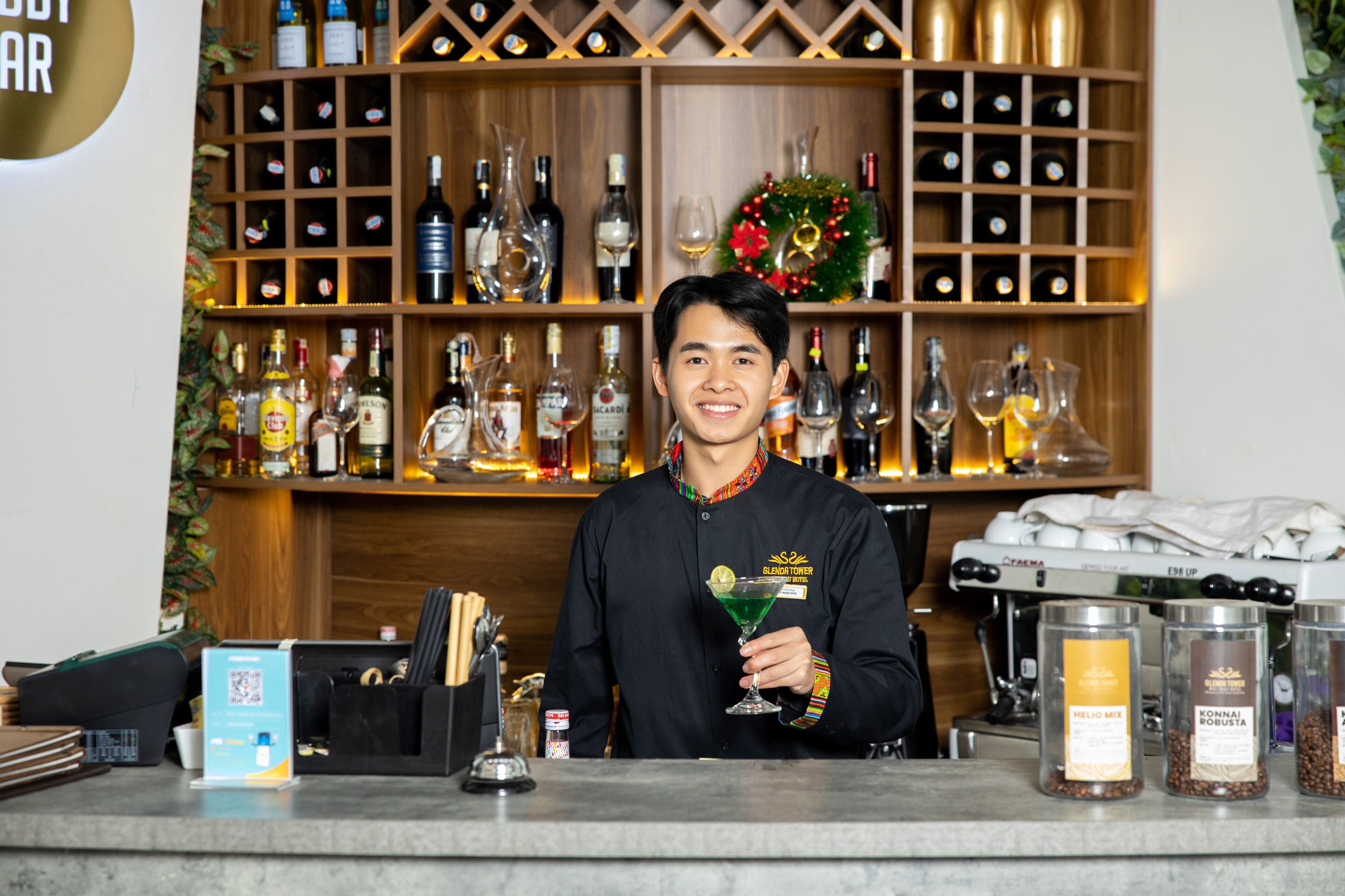 Eine Bar mit Regalen voller Flaschen und Gläsern, einer Kaffeemaschine und einer Person, die ein Cocktailglas hält.