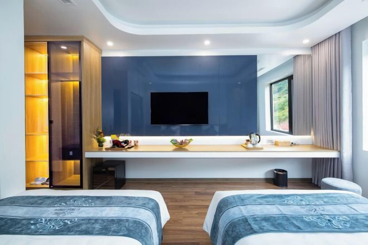 Una habitación de hotel moderna con un televisor de pantalla plana montado en la pared, un escritorio de madera con un frutero y dos camas con cabeceros a juego.