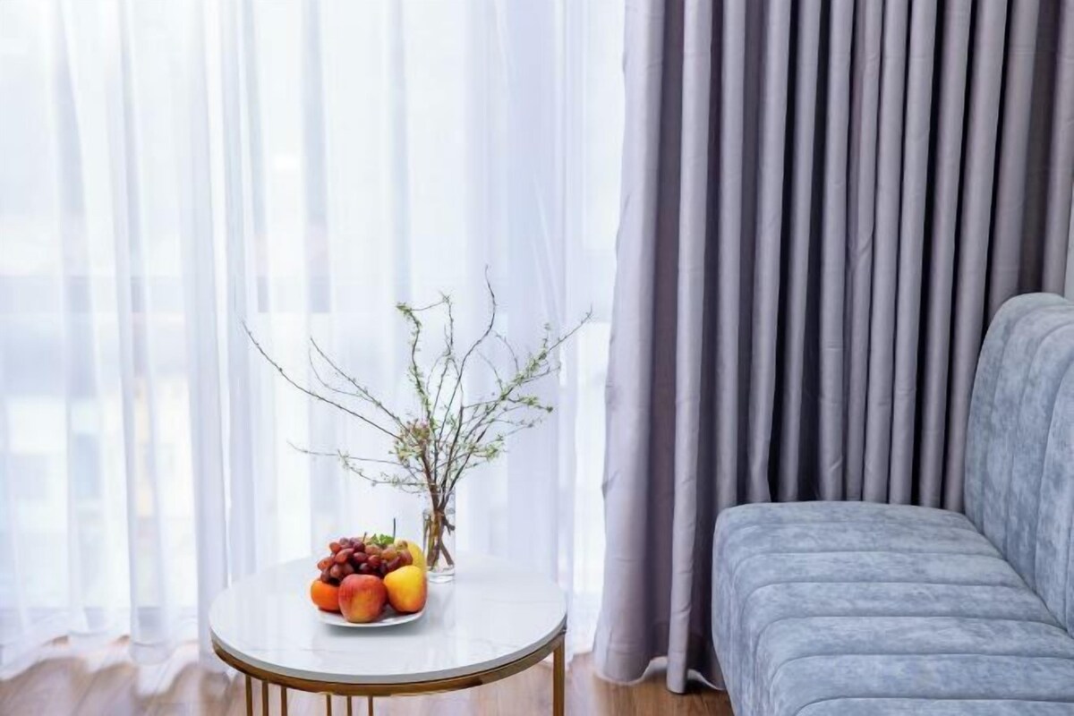 A room with a grey sofa, a round table with fruits and a vase, and sheer curtains.