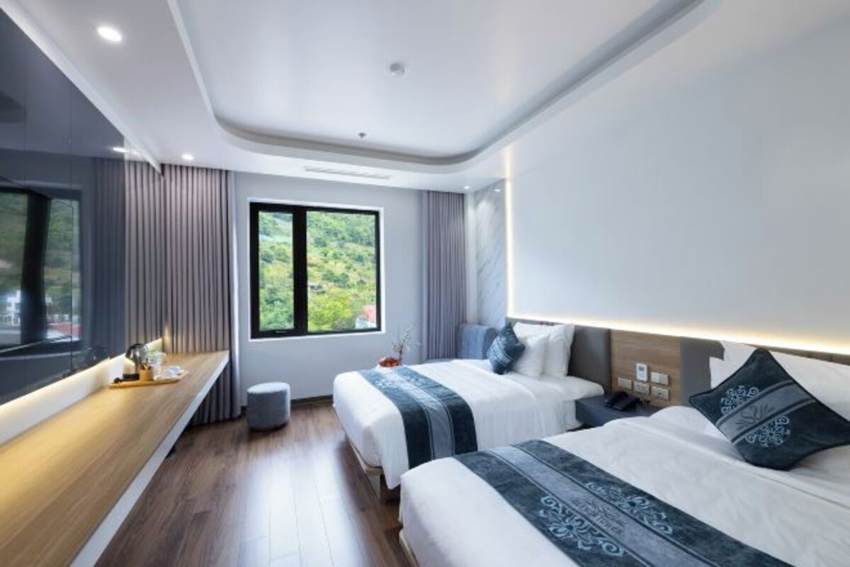 A modern hotel room with two beds, a wooden bedside table, a large window with a view of greenery, and a built-in bench along the window.
