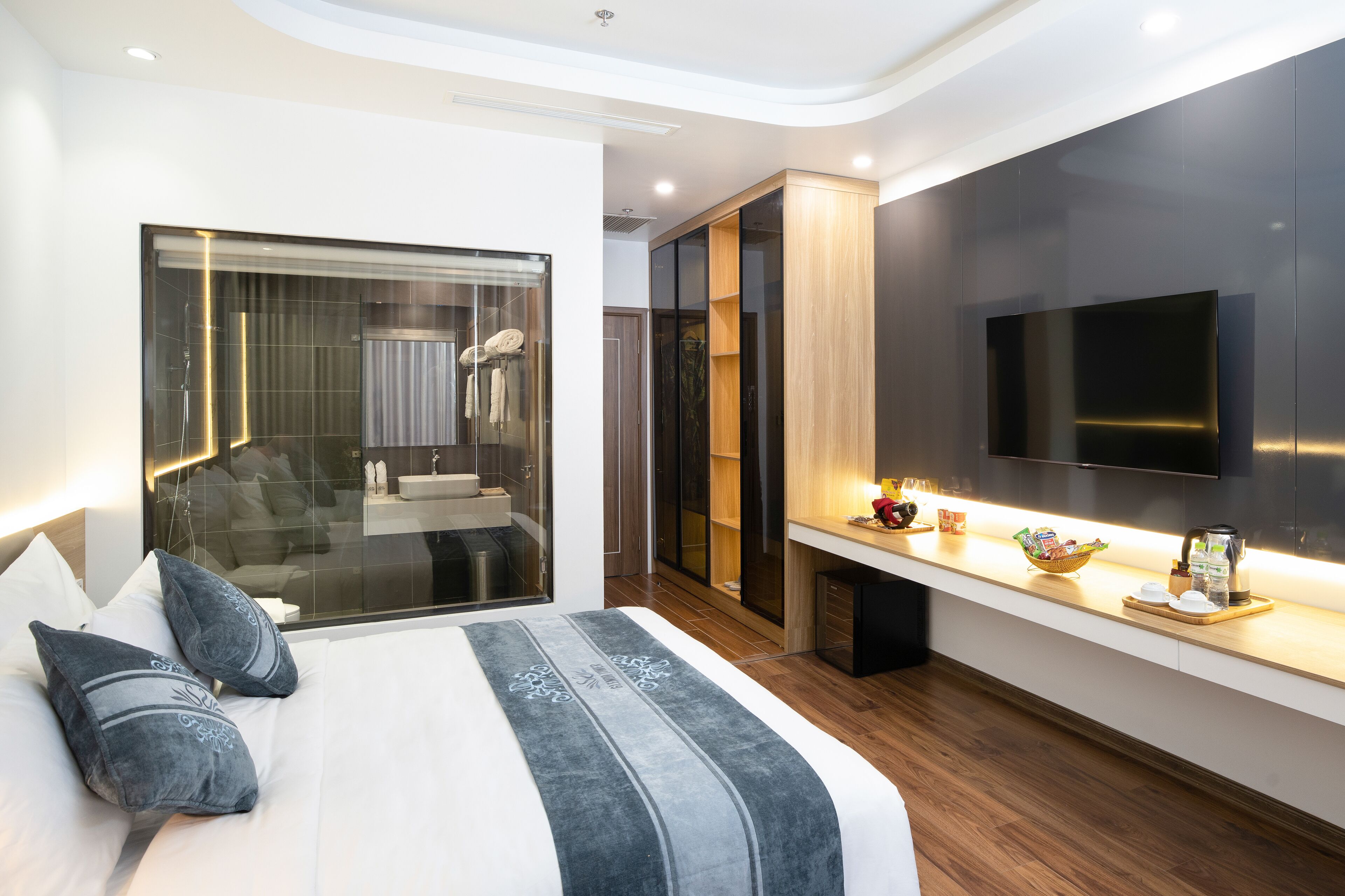 A modern hotel room with a large bed, a TV, a wooden floor, and a glass wall.